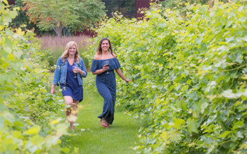 Two young women wearing dresses hold glasses of wine and walk through a green vineyard.