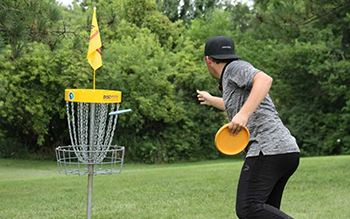 A young white man putts a disc into the basket at a green disc golf course