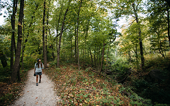 A woman in a light blue jacket hikes a gravel trail below green foliage.